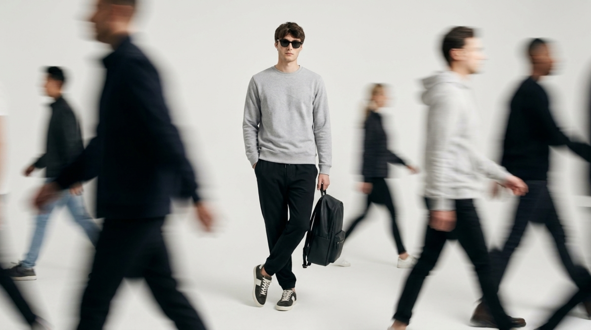 Minimalist fashion campaign featuring a man in a grey sweatshirt and black pants standing still while blurred people walk around him in motion against a clean white studio background.