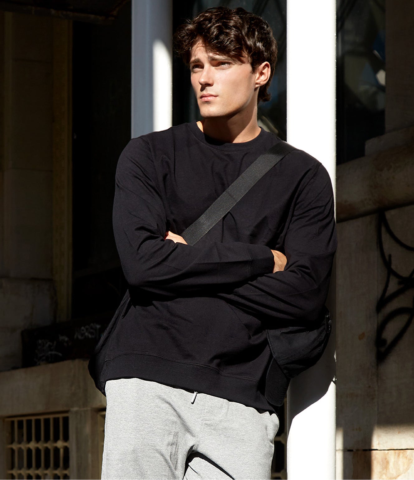Man wearing a black sweatshirt and gray pants standing against a building.