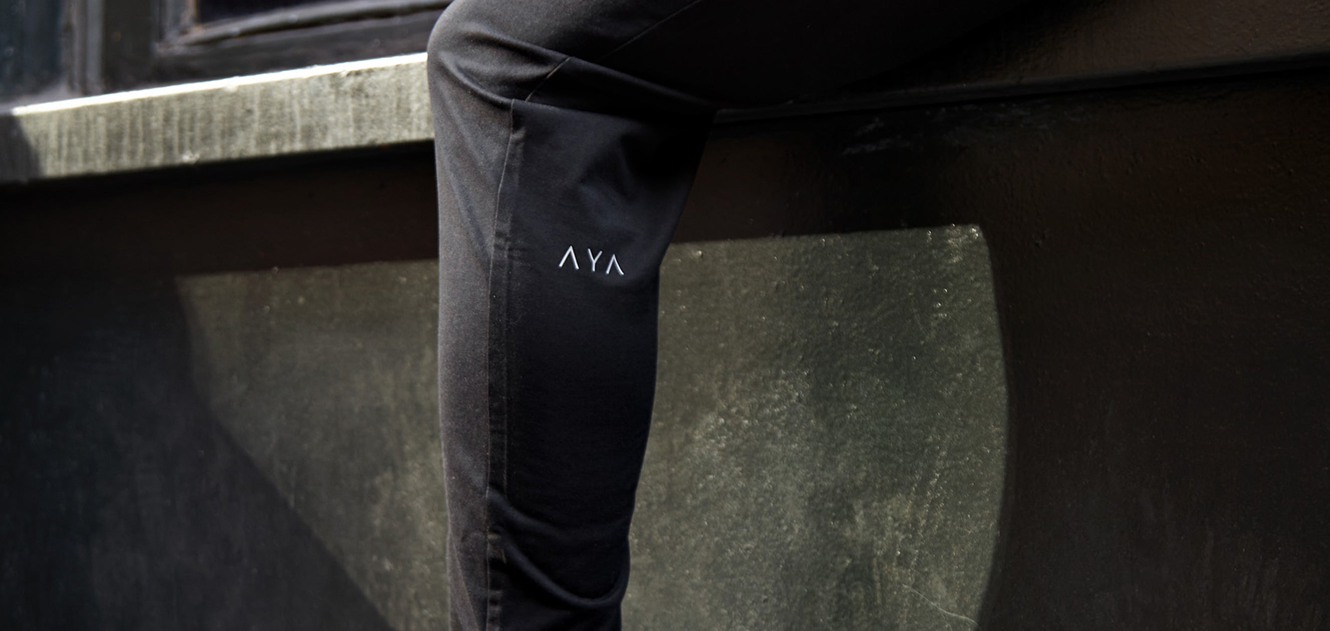Person wearing black jeans with a logo on the leg, sitting on a concrete surface.