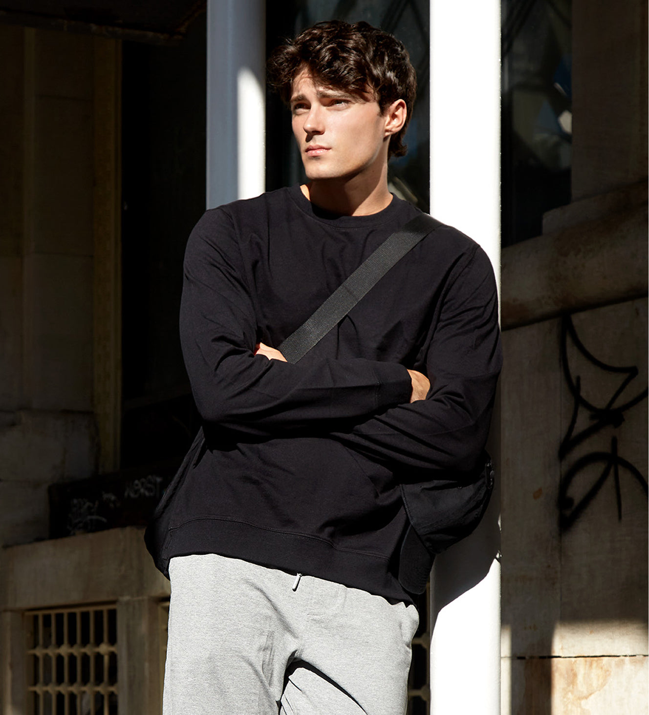 Man wearing a black sweater and gray pants standing against a stone wall.